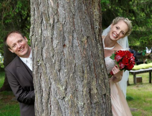 Ein Brautpaar schaut lächelnd hinter einem Baum hervor, die Braut hält einen Blumenstrauß mit roten Blumen