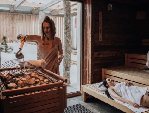 Mehrere Personen entspannen in Handtüchern in einer holzverkleideten Sauna mit einem Bild eines Waldes an der Wand