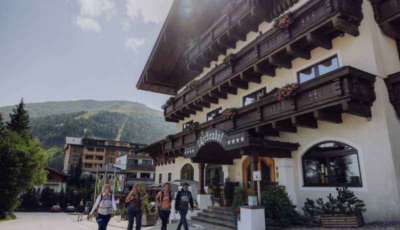 Außenansicht des Hotel Lärchenhof mit mehreren Personen, die zum Eingang gehen, und einer bergigen Landschaft im Hintergrund