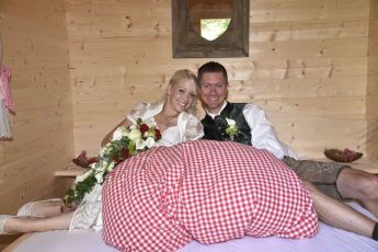 Ein Brautpaar in traditioneller Kleidung entspannt auf einem Bett mit rot-weiß karierten Kissen in einem rustikalen Raum