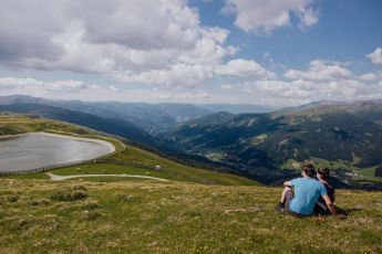 Ein Paar sitzt am Hang eines Hügels und blickt auf einen ruhigen Bergsee und das Tal